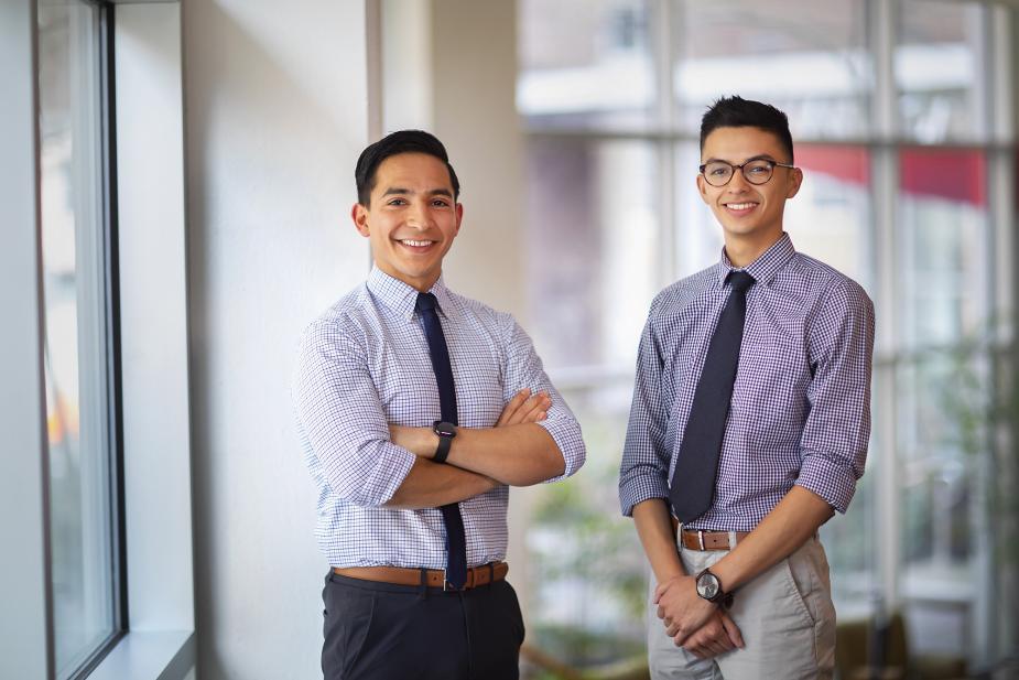  Two young men are standing side by side in a well-lit indoor setting. They are both wearing checkered shirts with rolled-up sleeves, dark ties, and dress pants. They are smiling and looking at the camera.