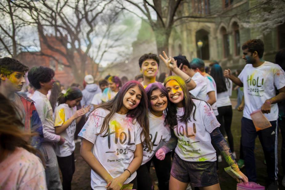 Four Penn students covered in colorful powder pose for a group picture during Holi fest. They are wearing matching Holi fest shirts.