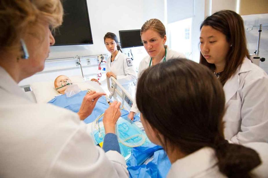 Two Penn Nursing instructors and three nursing students stand in scrubs around a hospital bed. There is a patient mannequin lying in the hospital bed and an instructor is demonstrating patient care for the watching students.