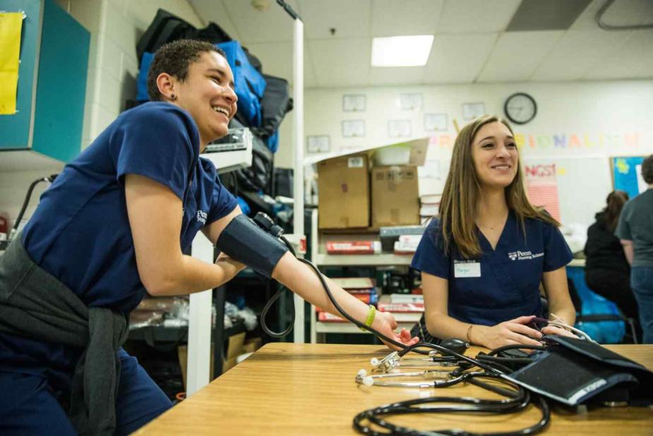 Penn nursing students are in a classroom practicing taking each other’s blood pressure. One student is smiling with her arm outstretched and a blood pressure cuff around her arm. The other is facing towards the front of the classroom with a stethoscope in her hands.