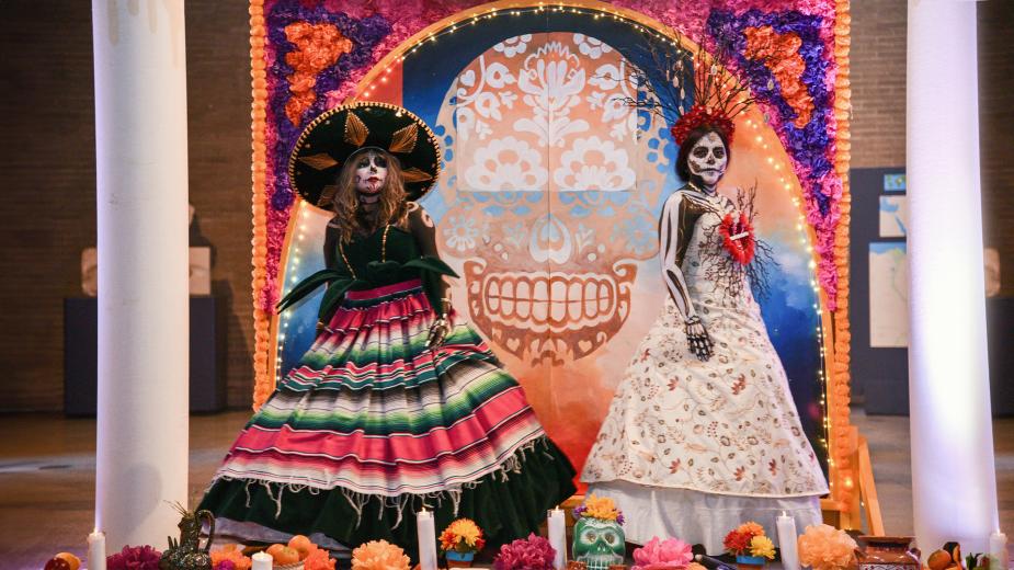 Two young women in traditional day of the dead clothing stand in front of a day of the dead altar. 