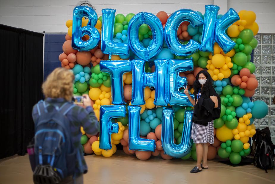 A person takes a photo of a woman posing in front of a 'BLOCK THE FLU' balloon display with colorful balloons in the background."