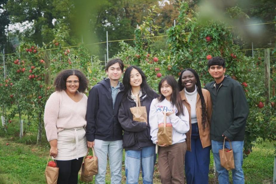 Six of Penn’s United Minorities Council students stand in a row in an apple orchard holding bags of apples they picked during a GIC event.