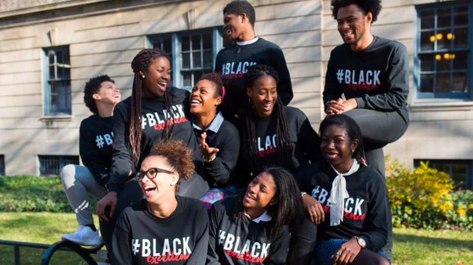 Group of 9 Makuu leadership students sit on a picnic table and converse outside. They are all wearing matching #BlackExcellence sweaters.