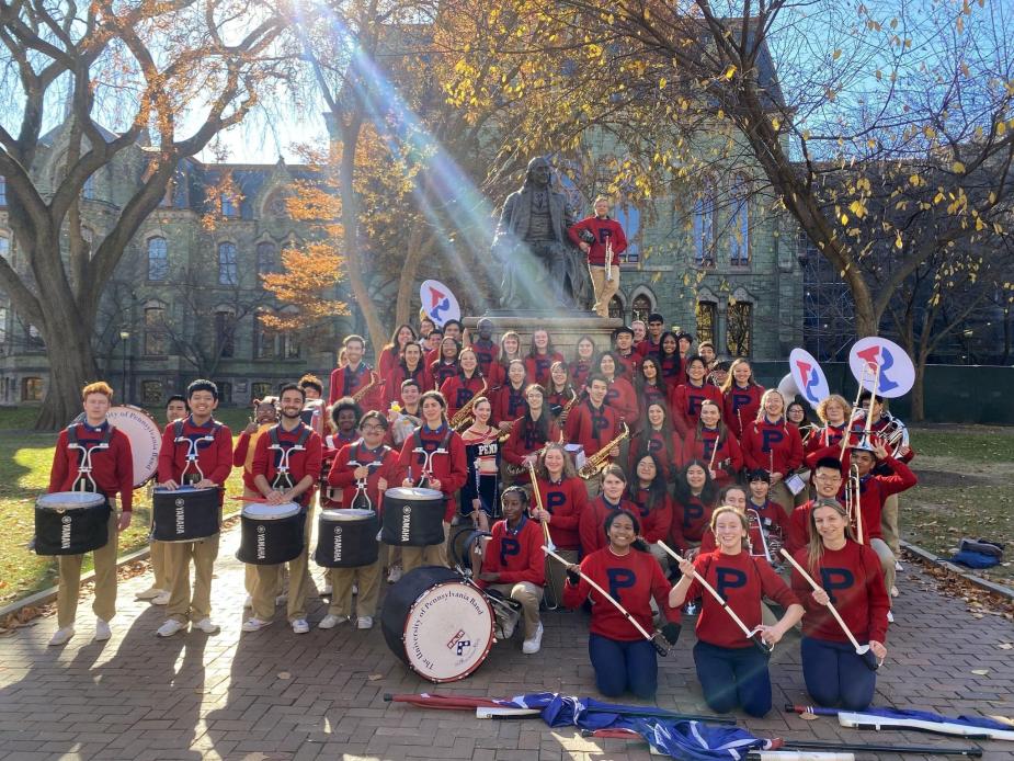 The Penn band is standing in front of the Ben Franklin statue on Penn's campus. They are wearing red sweaters with the Penn "P" and holding their instruments.