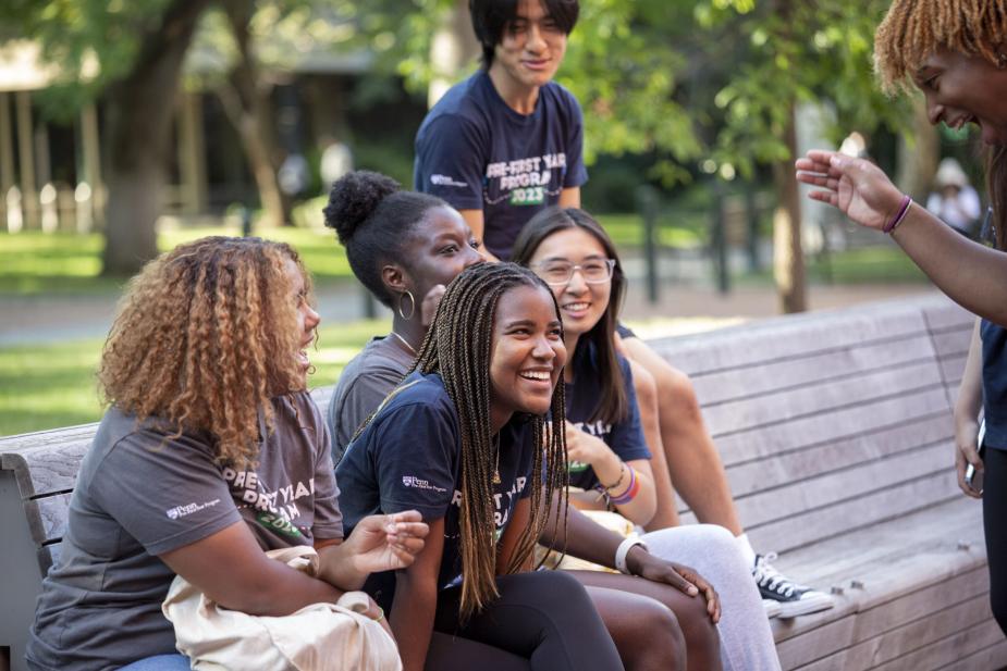 Penn students laugh and talk on an outdoor bench, wearing 'Penn First Plus' program shirts.