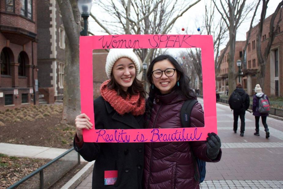 Two women outside pose for a picture with a square, pink picture frame that reads" Women Speak" at the top and then "Reality is Beautiful" at the bottom. 