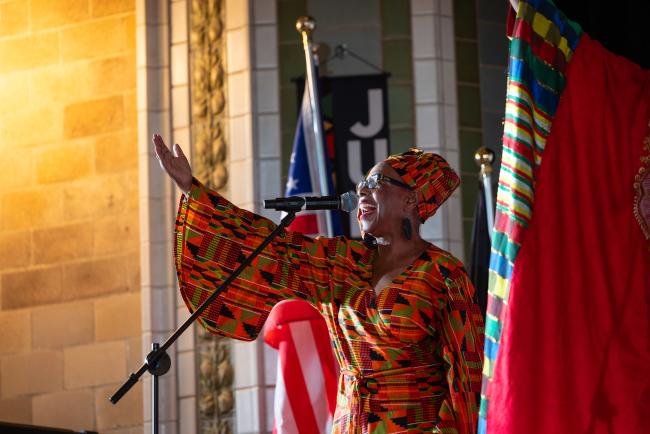 Woman in traditional African clothing sings at an event with her arm outstretched. 