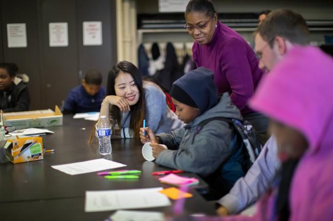 A group of people, including children and adults, are engaged in an indoor activity with papers, highlighters, and other supplies on a table. One adult is assisting a child who is writing.