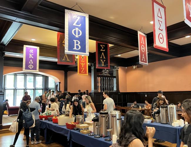 A room of Greek life students gathers for a meal. Above them hang different banners with Greek letters.