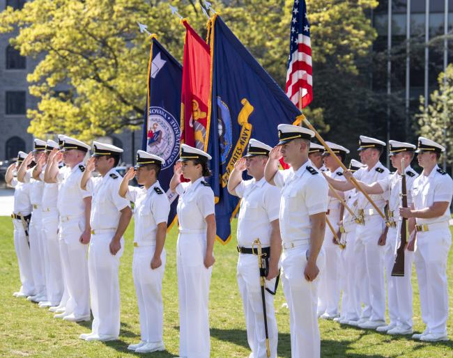 Two rows of NROTC students stand at attention on a lawn before several flags. 