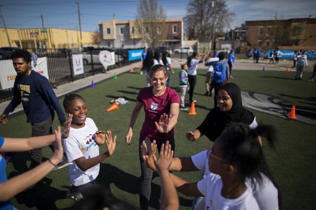 Penn student playing with a group of young children at an outdoor field. They are gathered in a circle and high fiving each other.