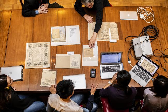 A large table is covered with different documents. Students and professors are looking at them together and taking notes on their laptops. 