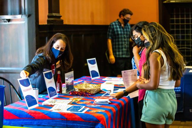  Students are at a Penn event table, checking out information on the event. They're talking to a staff member wearing a Penn mask. The table is covered with colorful materials and brochures.