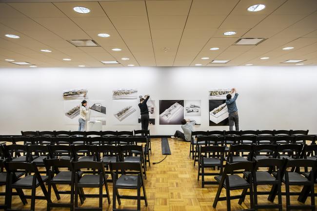 Three design students adhering their work to a wall in front of several chairs