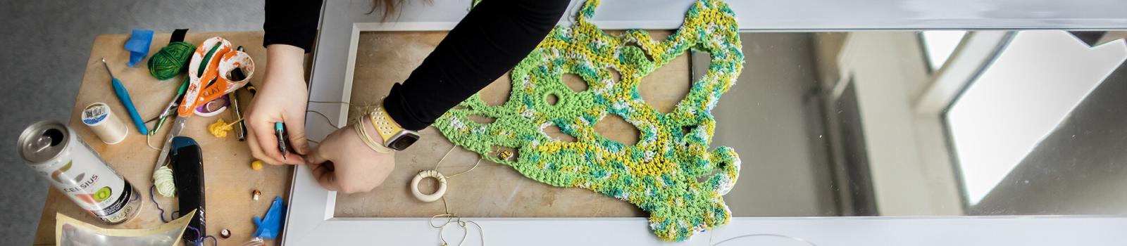 A large table is covered in scraps of fabric and an individual hovers over them creating a green, knitted artwork. They are working in Penn’s fabrication lab.