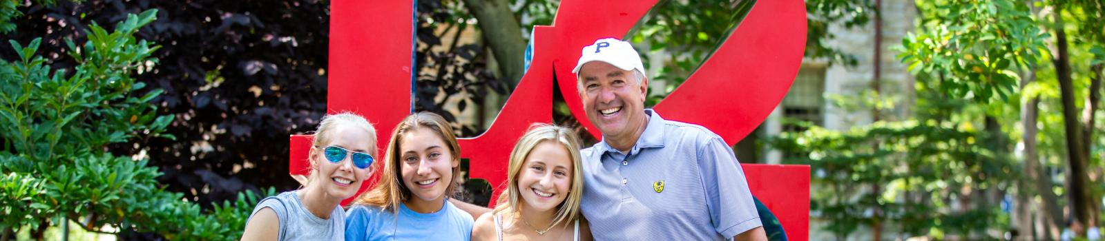 A family of four is posing in front of the LOVE sculpture at the University of Pennsylvania. They’re all smiling, with the parents on the ends and two young women in the middle.