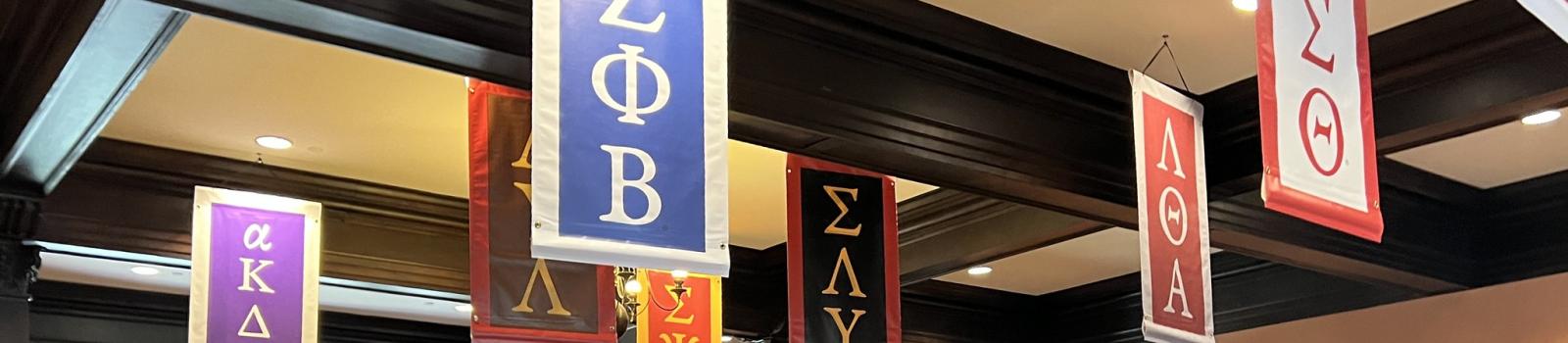 A room of Greek life students gathers for a meal. Above them hang different banners with Greek letters.
