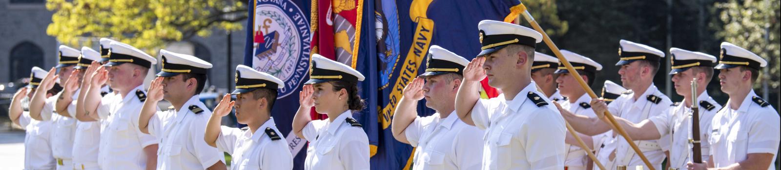 Two rows of NROTC students stand at attention on a lawn before several flags. 