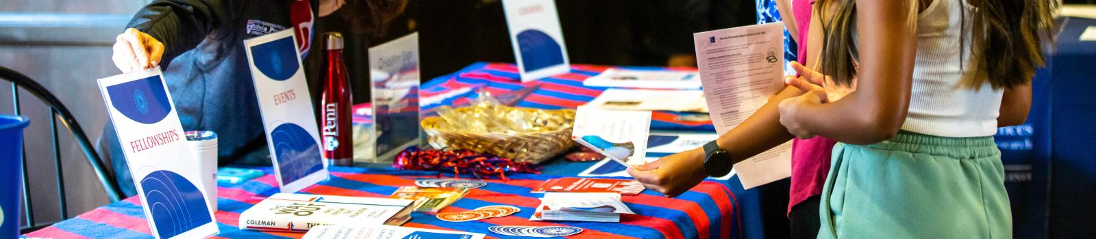  Students are at a Penn event table, checking out information on the event. They're talking to a staff member wearing a Penn mask. The table is covered with colorful materials and brochures.